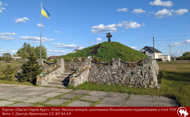 Фото курган-могили влітку. Зелений курган з козацьким хрестом на горі. До нього ведуть невисокі сходи. Навколо кургану змурований кам'яний парапет.